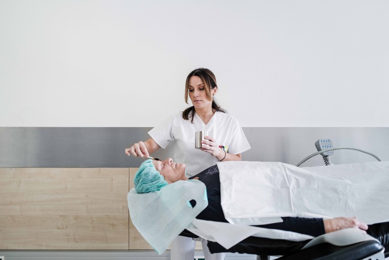 : Eine Patientin liegt auf einer Liege mit geschlossenen Augen und einer OP Haube. Eine Mitarbeiterin reinigt und desinfiziert das Gesicht mit einem gro&szlig;en Wattest&auml;bchen / &copy; Till Findl